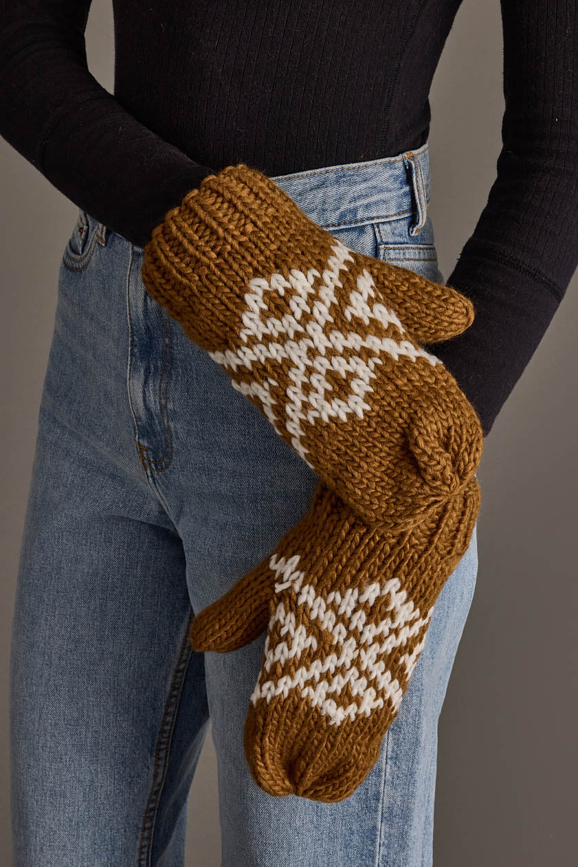 A person wearing blue jeans and a black long-sleeve top holds a pair of Camel & White Patterned Mittens from Panache Apparel Collection. The chunky mittens feature a white geometric design and appear warm and cozy.