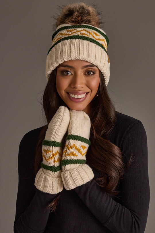 A woman smiles at the camera against a plain background, wearing Cream, Green & Gold Patterned Mittens by Panache Apparel Collection, along with a cream acrylic knit hat featuring a pom-pom.