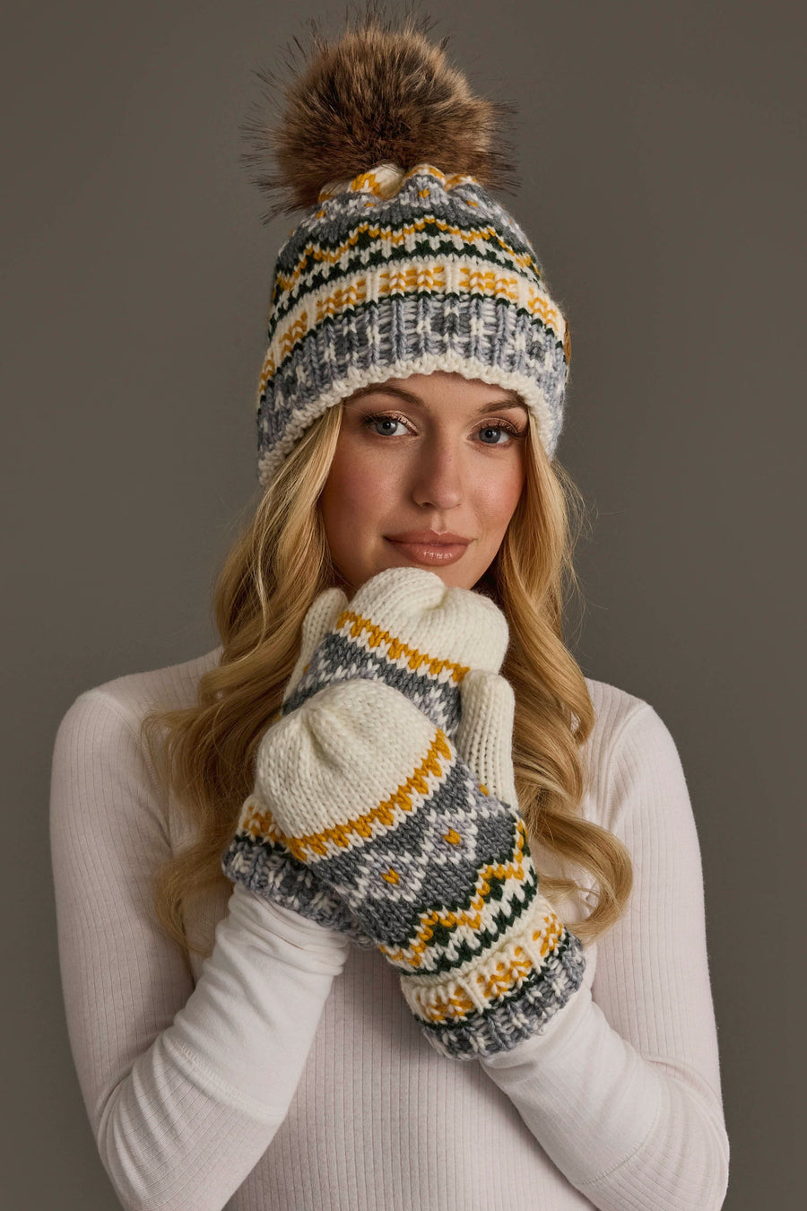 A woman wearing a white sweater and matching patterned mittens smiles gently as she poses against a plain gray background, accessorized with the Ivory, Green, Gray & Gold Patterned Pom Hat from Panache Apparel Collection.