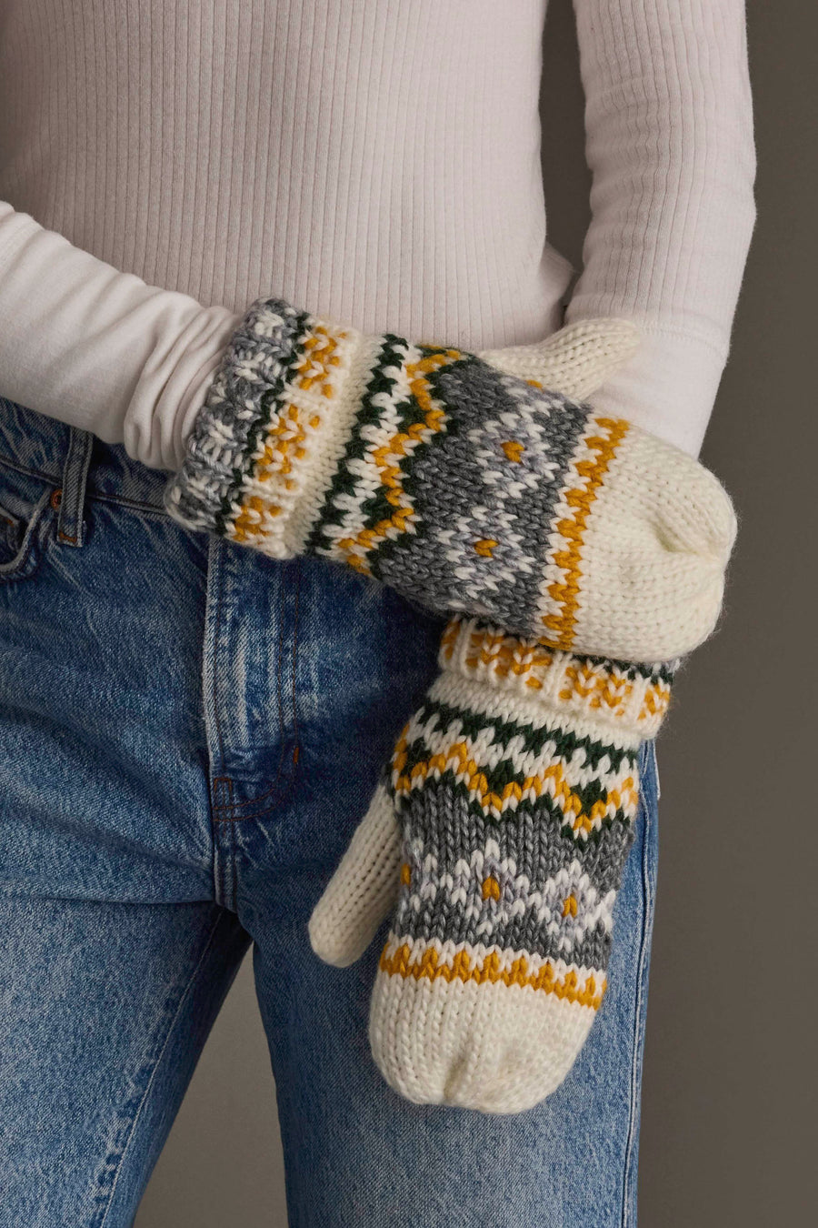 A person wearing blue jeans and a white ribbed long-sleeve shirt holds hands adorned with Panache Apparel Collection’s Ivory, Green, Gray & Gold Patterned Mittens.