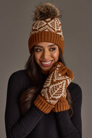 A woman wearing the Panache Apparel Collection Camel & Beige Patterned Knit Pom Hat smiles as she holds her matching mittens with white geometric patterns to her face, set against a neutral background.