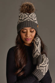 A woman models the Panache Apparel Collection’s Gray & Beige Patterned Knit Pom Hat, featuring a playful pom-pom, paired with matching mittens against a plain dark background.
