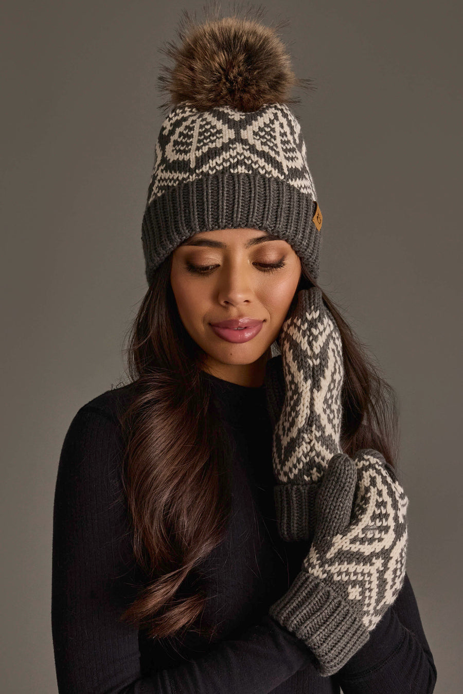 A woman models the Panache Apparel Collection’s Gray & Beige Patterned Knit Pom Hat, featuring a playful pom-pom, paired with matching mittens against a plain dark background.