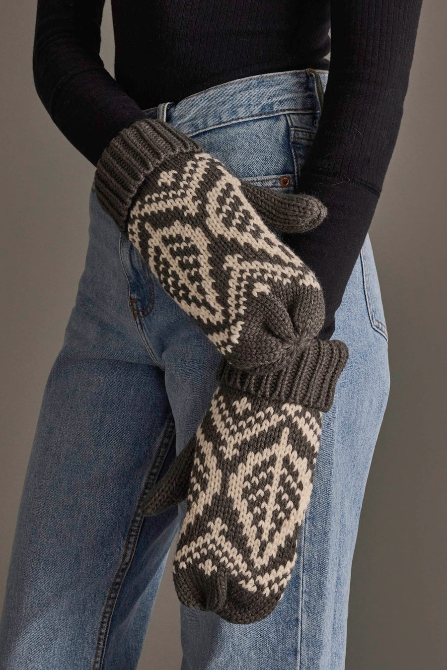 A person wearing blue jeans, a black long-sleeve top, and the Panache Apparel Collection’s Gray & Beige Patterned Knit Mittens stands with their arms crossed.