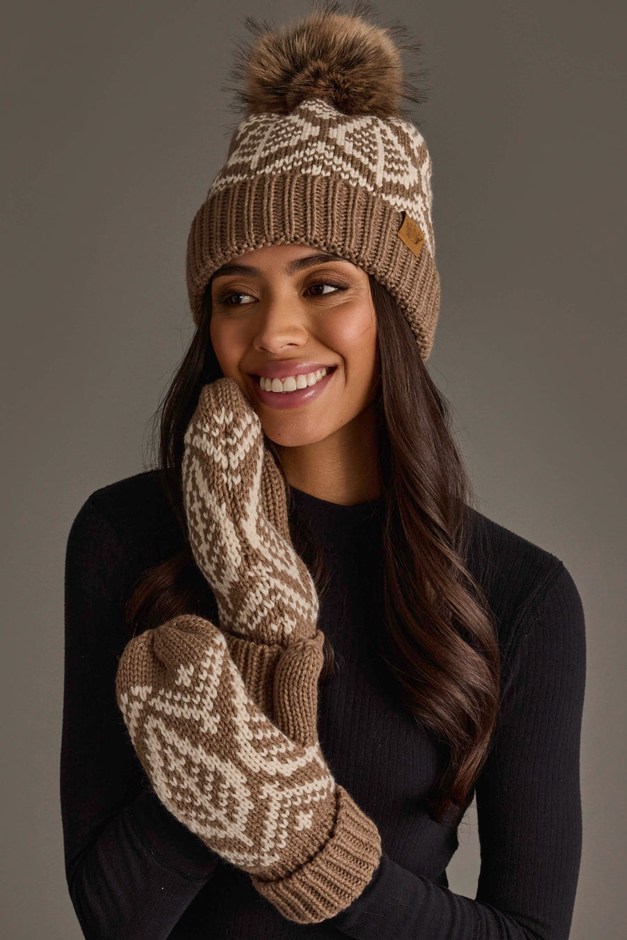 A woman with long, dark hair is smiling and looking to the side while wearing the Panache Apparel Collection Taupe & Beige Patterned Knit Pom Hat. She is also wearing matching mittens and a black top against a plain gray background.