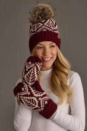 A smiling woman in a white sweater wears the Panache Apparel Collection Wine & Beige Patterned Knit Pom Hat, paired with matching maroon and white patterned mittens, standing against a gray background.