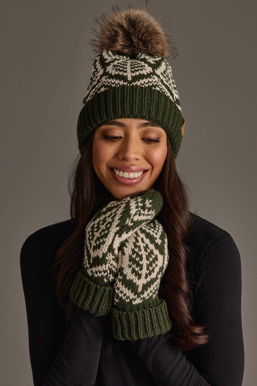 A woman wearing the Olive & Beige Patterned Knit Pom Hat from Panache Apparel Collection and matching green and white mittens smiles with her hands near her face against a neutral background.