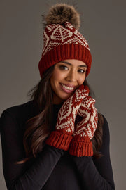 A woman smiles at the camera against a plain gray background, wearing the Panache Apparel Collection Rust & Beige Patterned Knit Pom Hat and matching patterned gloves.