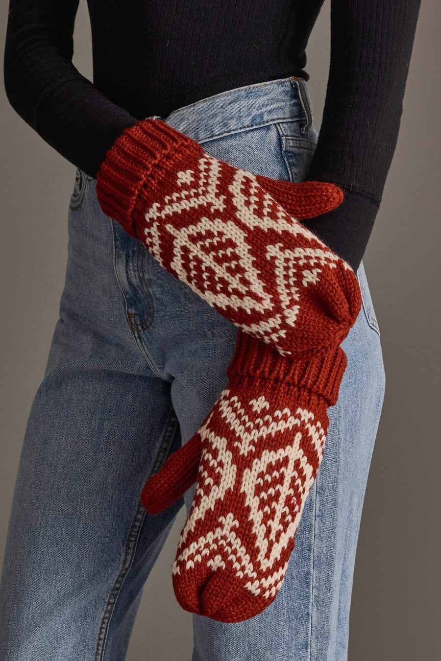 A person wearing blue jeans and a black sweater displays the Panache Apparel Collection’s Rust & Beige Patterned Knit Mittens.