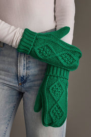 A person in a white ribbed long-sleeve shirt and light blue jeans holds up a pair of Panache Apparel Collection Bright Classic Green Cable Knit Mittens featuring a chunky textured diamond pattern.