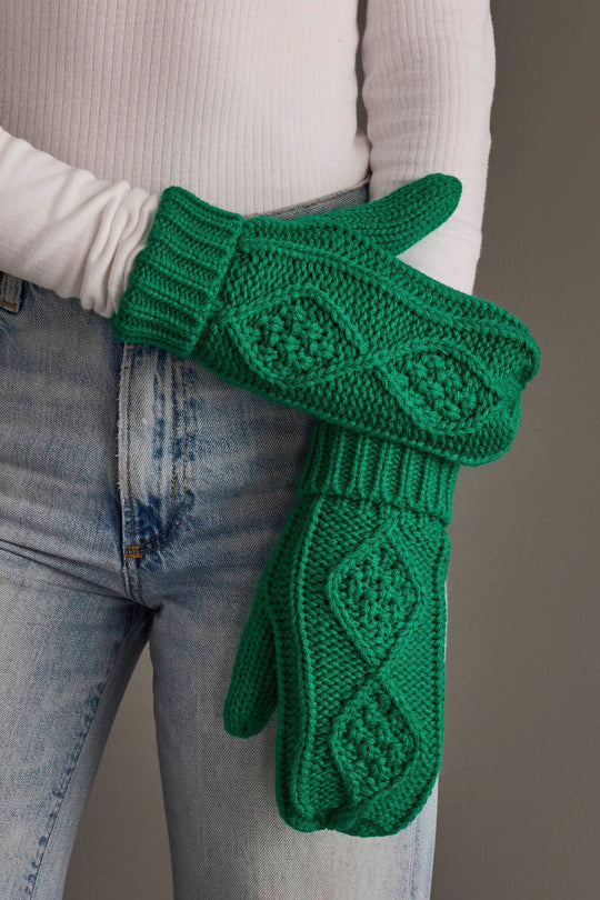 A person in a white ribbed long-sleeve shirt and light blue jeans holds up a pair of Panache Apparel Collection Bright Classic Green Cable Knit Mittens featuring a chunky textured diamond pattern.