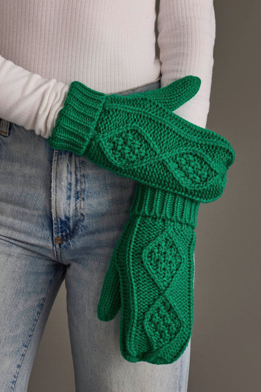 A person in a white ribbed long-sleeve shirt and light blue jeans holds up a pair of Panache Apparel Collection Bright Classic Green Cable Knit Mittens featuring a chunky textured diamond pattern.