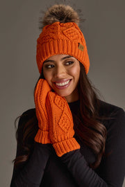 A woman smiles while wearing the Orange Cable Knit Pom Hat from Panache Apparel Collection, paired with matching orange knit gloves and a black long-sleeve top, posed against a plain gray background.
