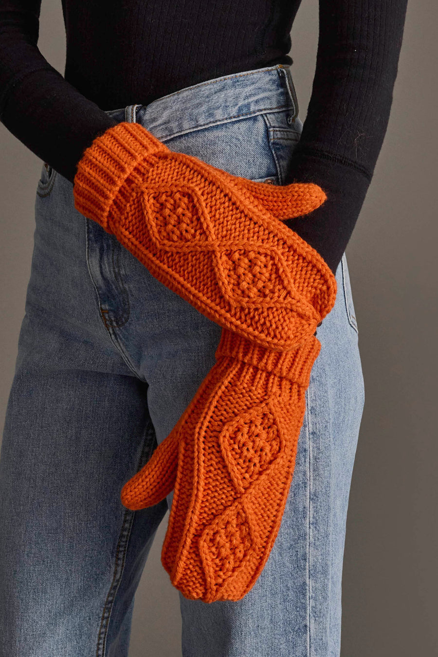 A person dressed in high-waisted blue jeans and a black long-sleeve top holds out the Orange Cable Knit Mittens by Panache Apparel Collection, which feature bright orange coloring with diamond and textured knit patterns. The background is a neutral gray.