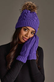 A woman wears the Panache Apparel Collection Purple Cable Knit Pom Hat, featuring a faux fur pom-pom, along with matching purple knit mittens. She holds her hands to her face and smiles gently.