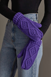 A person wearing blue jeans and a black top holds a pair of Panache Apparel Collection Purple Cable Knit Mittens featuring textured diamond patterns.
