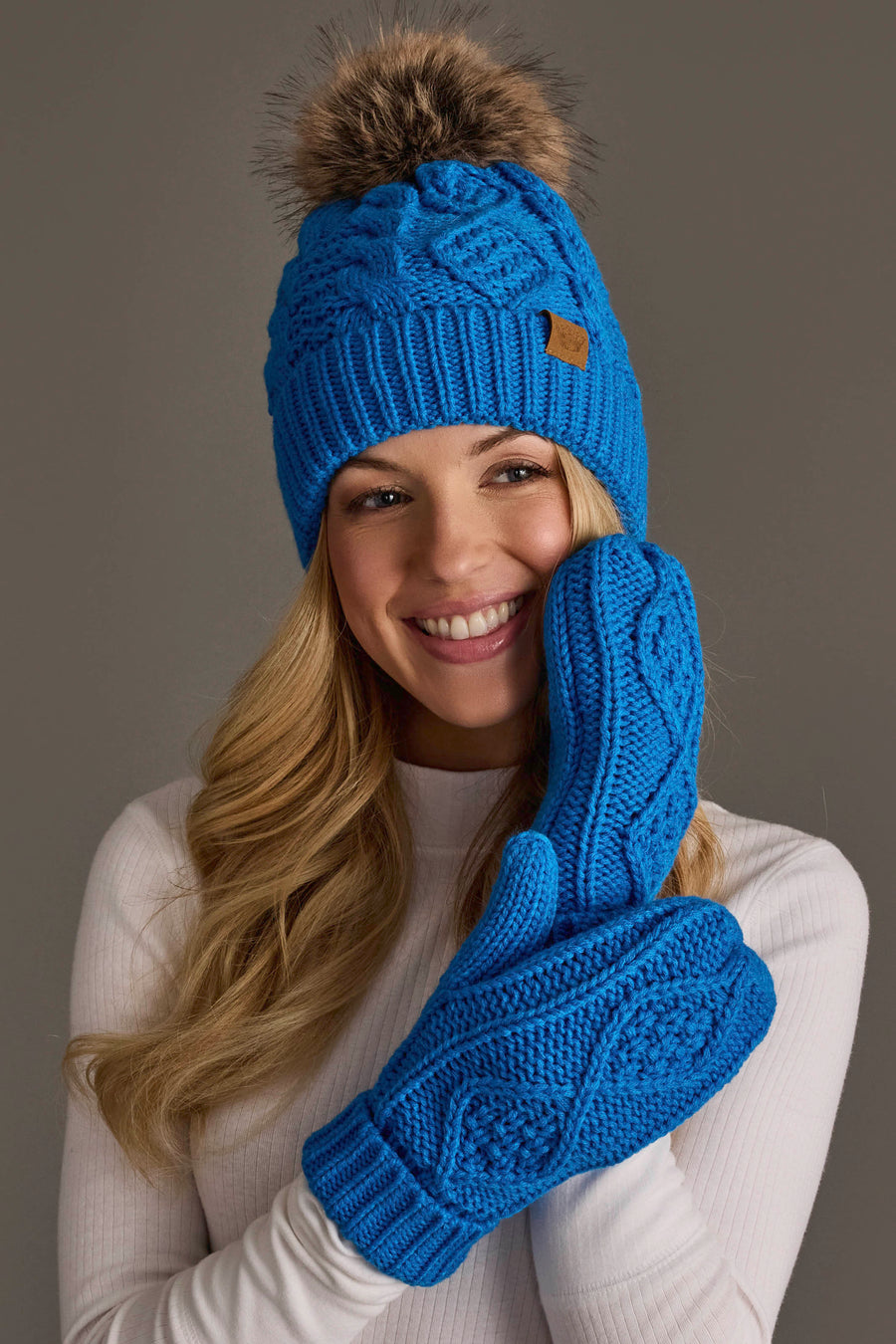 A woman in a white sweater smiles while wearing the Panache Apparel Collection’s Blue Cable Knit Pom Hat, styled with matching blue knit mittens, as she poses against a plain, dark background.