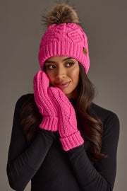 A woman, dressed in a black long-sleeve top, holds her hands to her face while wearing the Panache Apparel Collection’s Bright Pink Cable Knit Pom Hat with a fur pompom and matching pink mittens, set against a plain gray background.