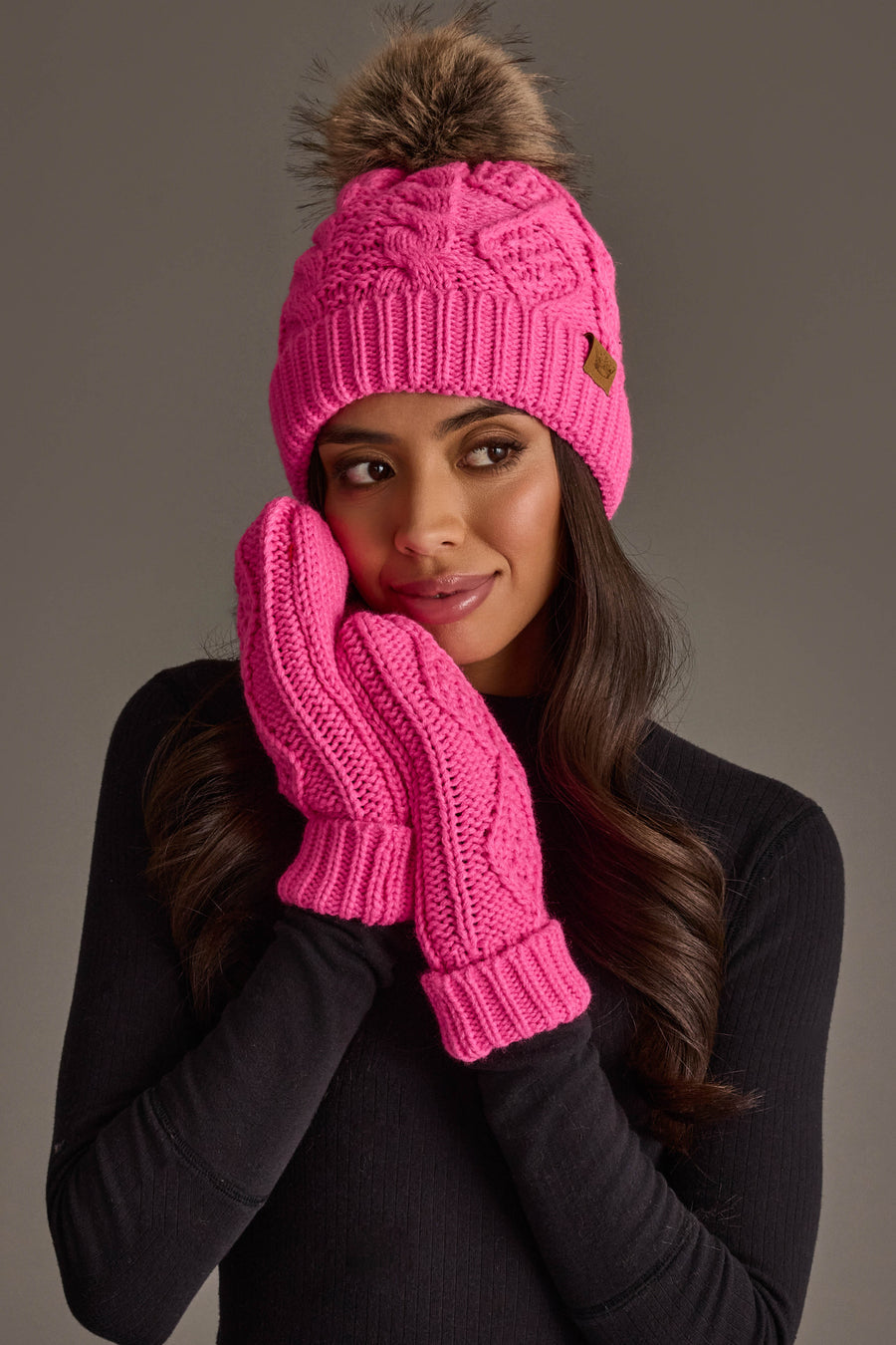A woman, dressed in a black long-sleeve top, holds her hands to her face while wearing the Panache Apparel Collection’s Bright Pink Cable Knit Pom Hat with a fur pompom and matching pink mittens, set against a plain gray background.