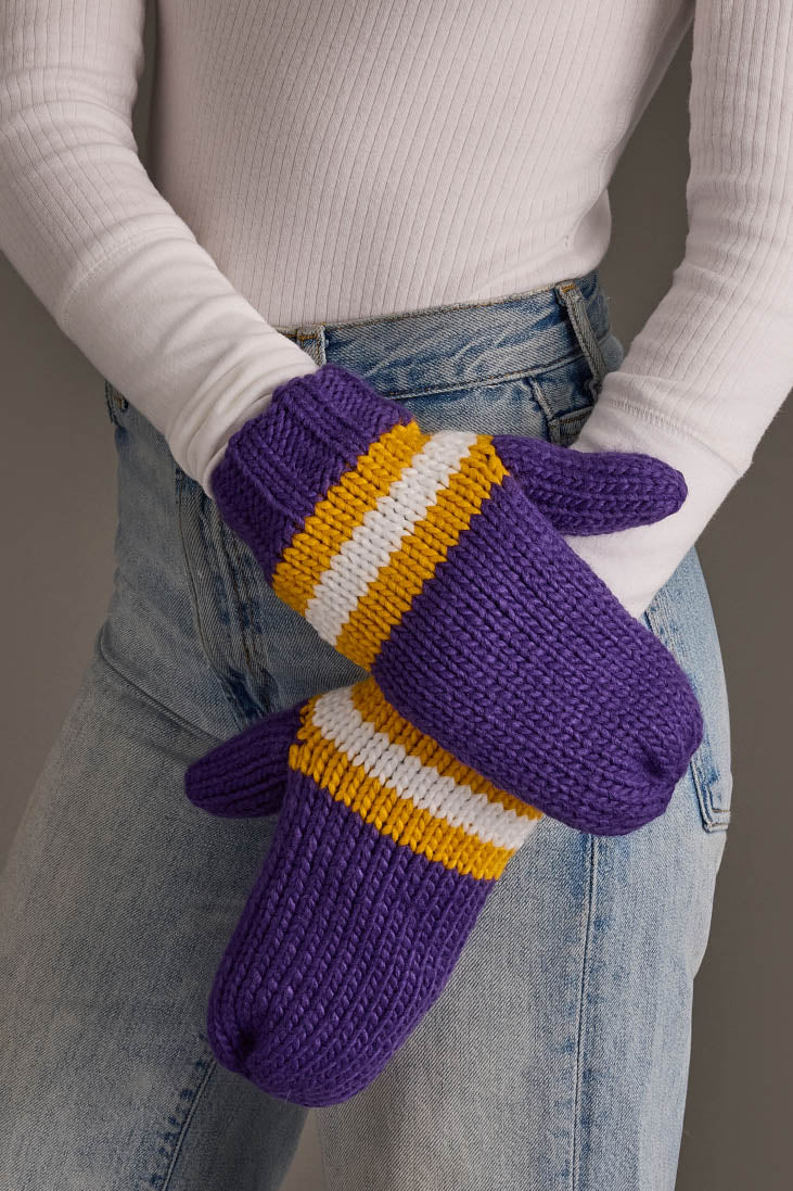 A person in a white ribbed turtleneck and blue jeans stands with arms crossed, wearing the Panache Apparel Collection’s Purple, Gold & White Knit Mittens.