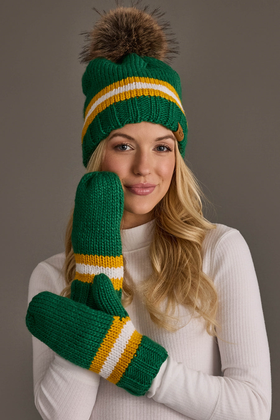 A smiling woman in a white turtleneck wears the Panache Apparel Collection Green, Gold & White Knit Pom Hat, posing against a plain background.