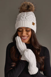 A woman smiles with her eyes closed, wearing the Panache Apparel Collection White Cable Knit Pom Hat and matching white mittens, dressed in a black top against a gray background.