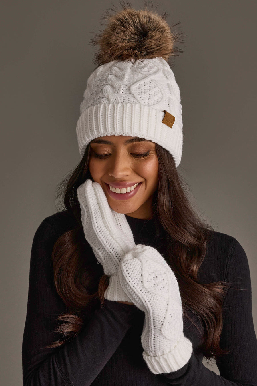 A woman smiles with her eyes closed, wearing the Panache Apparel Collection White Cable Knit Pom Hat and matching white mittens, dressed in a black top against a gray background.