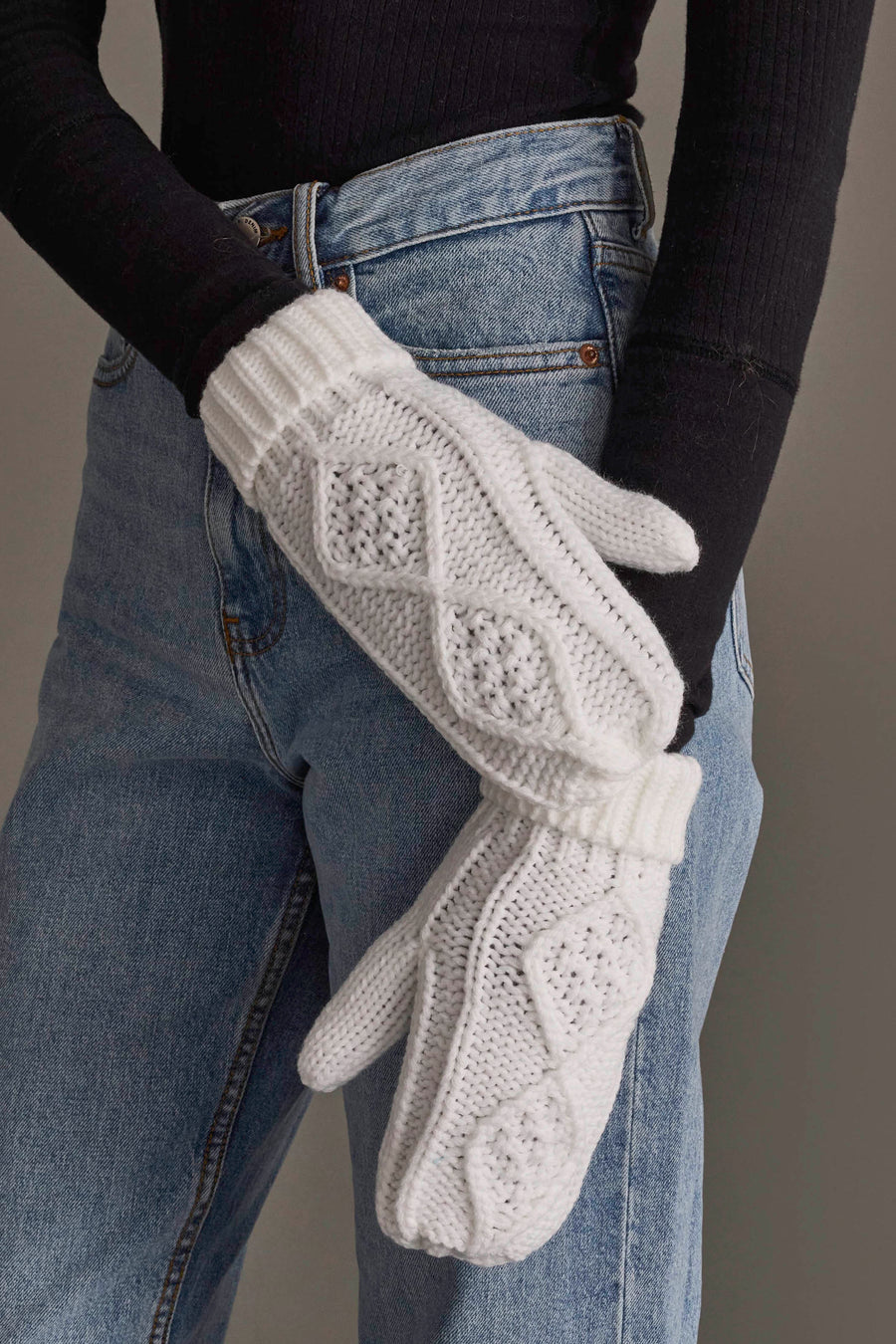 A person wearing blue jeans, a black long-sleeve top, and White Cable Knit Mittens from Panache Apparel Collection stands with arms crossed.