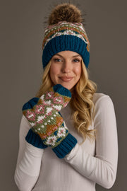 A woman in a white shirt smiles as she holds her hands near her face, wearing the Panache Apparel Collection Blue, Green, Pink & Brown Patterned Pom Hat and matching mittens, set against a gray background.