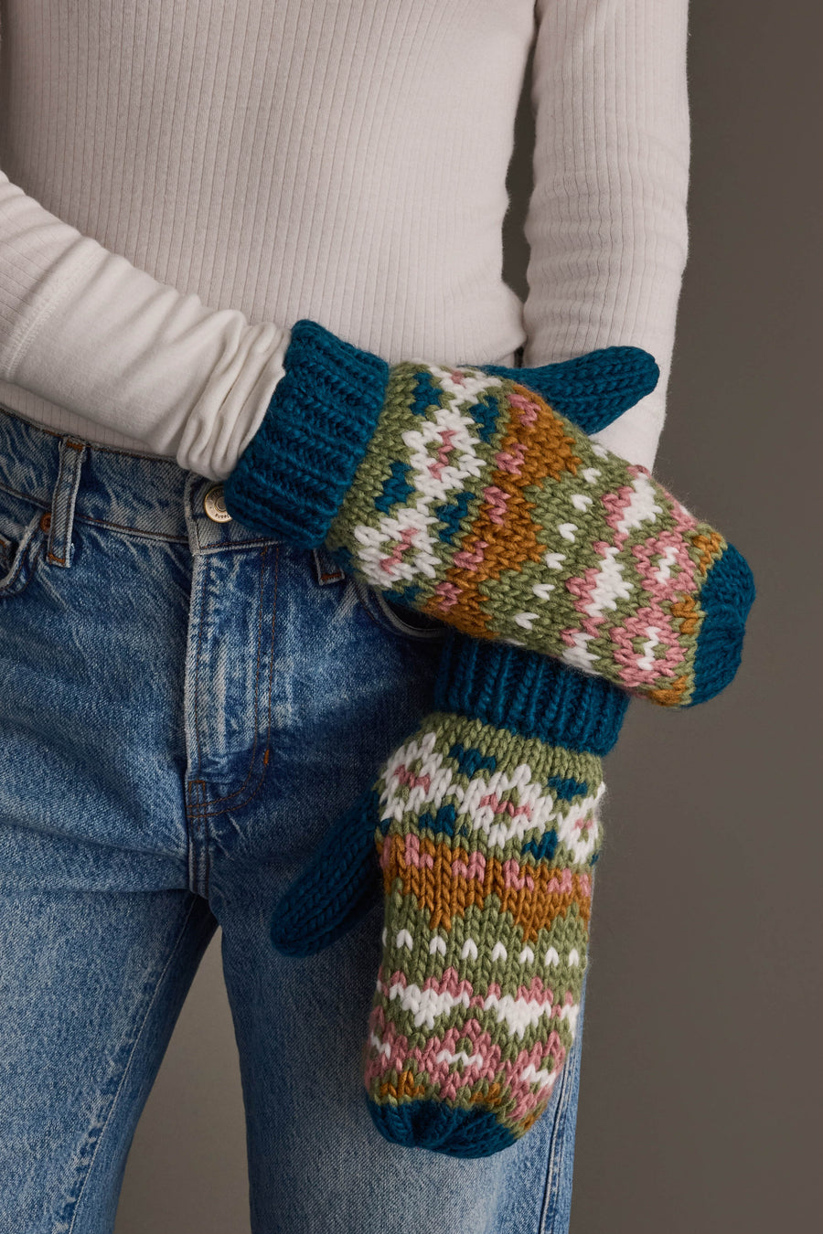 A person in a white long-sleeve shirt and blue jeans showcases the Panache Apparel Collection’s Blue, Green, Pink & Brown Patterned Mittens, chunky hand-knit mittens featuring a colorful fair isle design and ribbed cuffs.