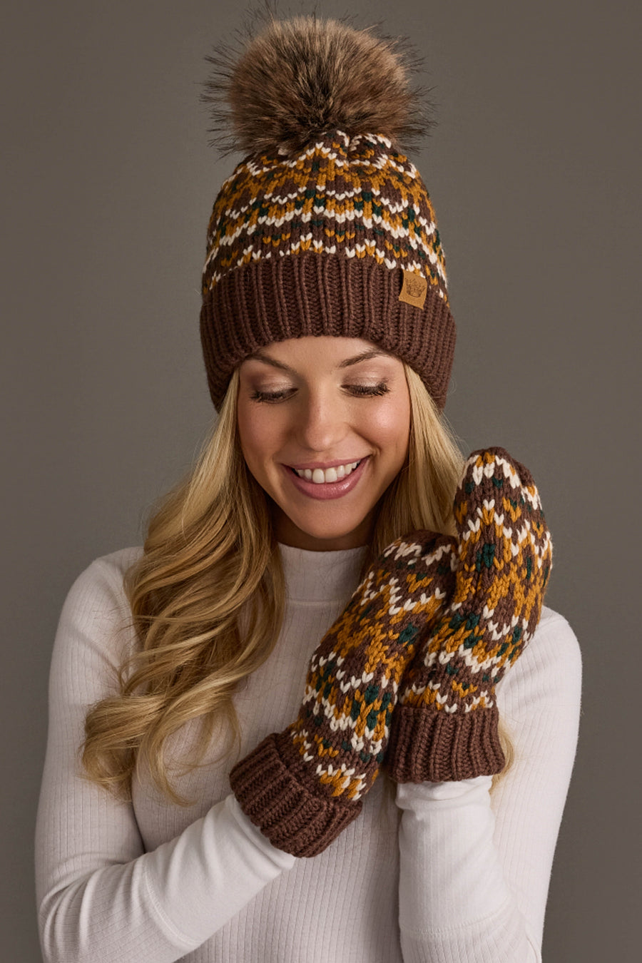 A smiling woman wears the Panache Apparel Collection’s Brown, Mustard & Cream Patterned Pom Hat along with matching mittens and a white sweater, standing against a neutral background.