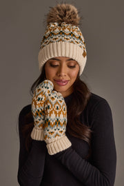 A woman with long dark hair stands against a plain gray background, wearing the Panache Apparel Collection’s Beige, Mustard & Green Patterned Pom Hat with matching mittens and a black long-sleeve top.