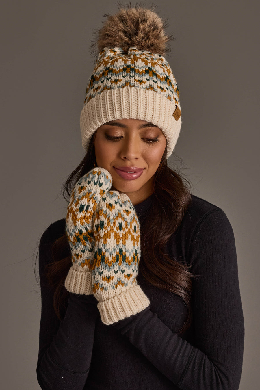 A woman with long dark hair stands against a plain gray background, wearing the Panache Apparel Collection’s Beige, Mustard & Green Patterned Pom Hat with matching mittens and a black long-sleeve top.