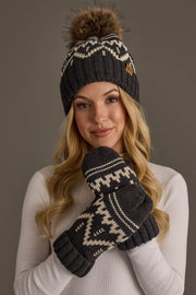 A woman with long blonde hair wears the Charcoal & Beige Patterned Pom Hat from Panache Apparel Collection along with matching mittens, both featuring a dark gray and white design. She is dressed in a white top and smiles softly at the camera.
