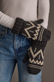 A person in blue jeans and a white ribbed sweater holds the Charcoal & Beige Patterned Mittens by Panache Apparel Collection, featuring geometric designs.