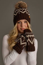 A woman wearing a white sweater and the Panache Apparel Collection’s Chocolate Brown & Beige Patterned Pom Hat, featuring a furry pom-pom, along with matching mittens, poses against a plain gray background.