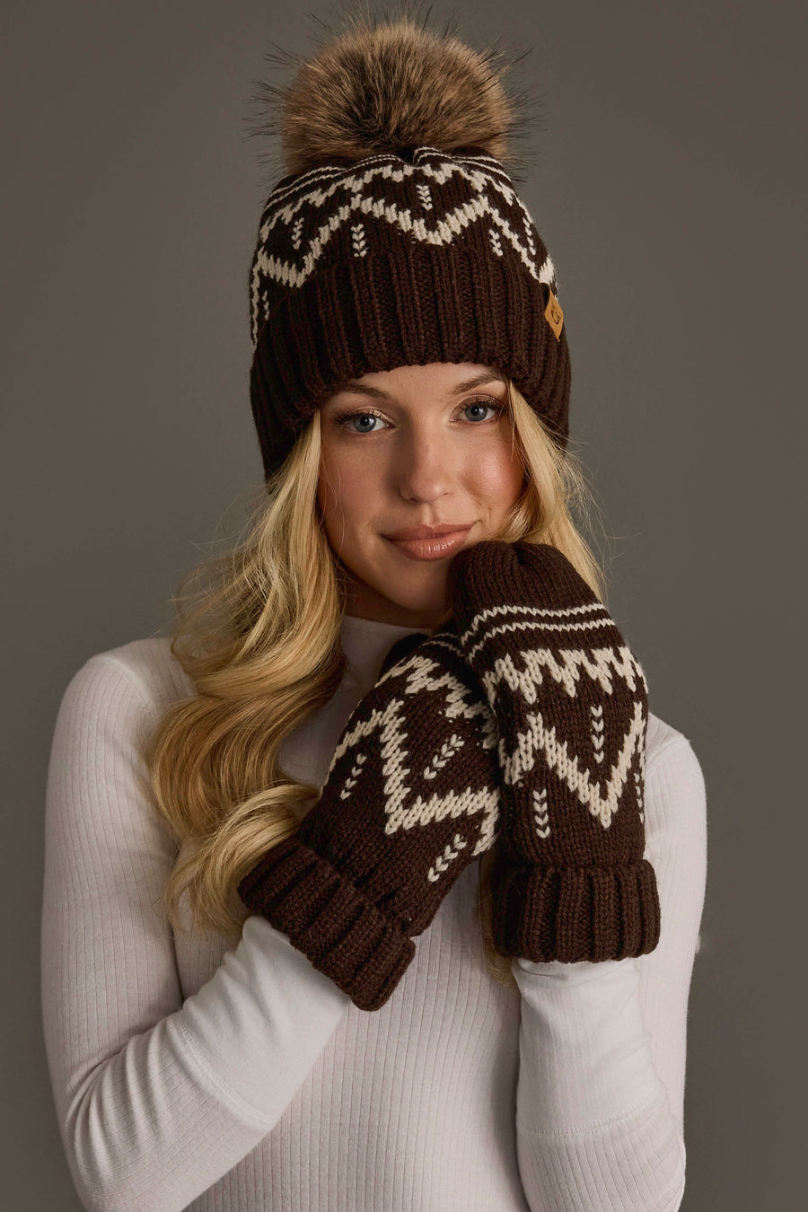 A woman wearing a white sweater and the Panache Apparel Collection’s Chocolate Brown & Beige Patterned Pom Hat, featuring a furry pom-pom, along with matching mittens, poses against a plain gray background.