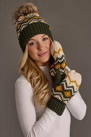 A person wearing the Panache Apparel Collection Olive, Cream & Mustard Patterned Pom Hat, paired with matching patterned mittens, smiles in a white long-sleeve shirt against a neutral background.