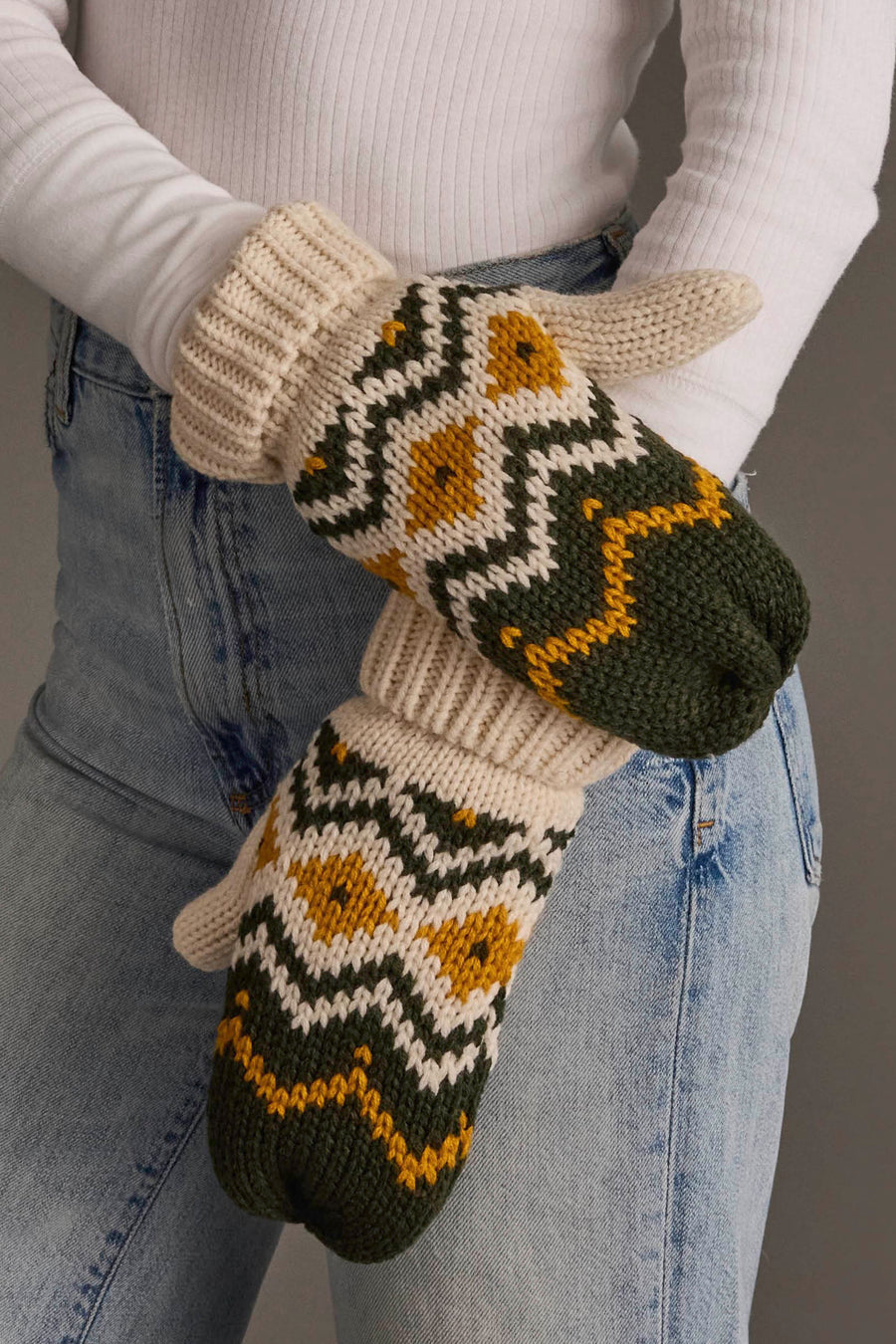 A person wearing blue jeans and a white ribbed long-sleeve shirt has their arms crossed at the waist, showcasing the Cream, Olive & Mustard Patterned Mittens from Panache Apparel Collection.