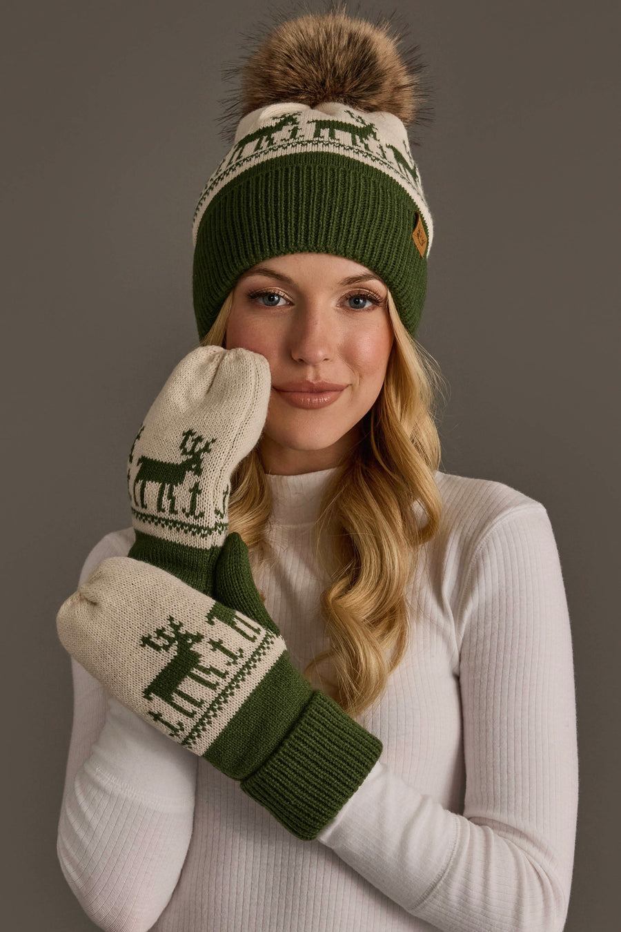 A person wears a Panache Apparel Collection Olive & Beige Deer Patterned Pom Hat and matching mittens, both featuring a reindeer pattern, beige and olive green fleece lining, and faux fur detail.