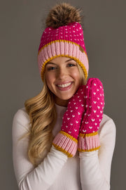 A smiling woman wears the Panache Apparel Collection Pink, White & Light Pink Heart Design Pom Hat, a pink knit hat with a fur pom-pom, paired with matching pink mittens with white dots and a light turtleneck top, standing against a plain background.