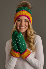 A smiling person in a white sweater wears the Panache Apparel Collection Green, White & Mustard Heart Design Pom Hat with matching mittens, featuring white heart details, while posing against a plain gray background.