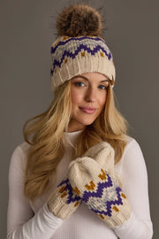 A woman in a white turtleneck models the Panache Apparel Collection Cream, Gold, Purple & Gray Patterned Pom Hat featuring a brown pom-pom and stylish zigzag details against a plain gray background.