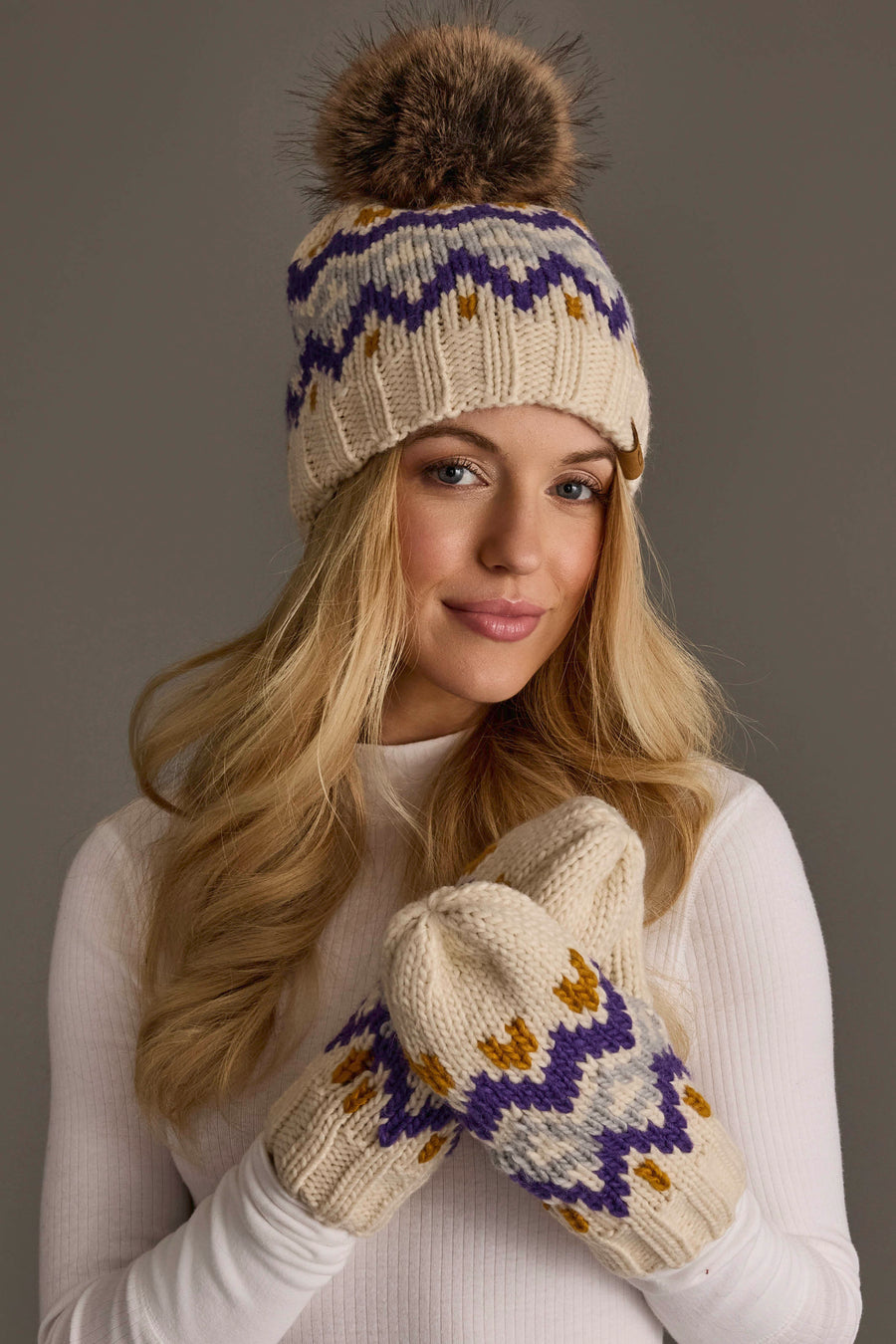 A woman in a white turtleneck models the Panache Apparel Collection Cream, Gold, Purple & Gray Patterned Pom Hat featuring a brown pom-pom and stylish zigzag details against a plain gray background.