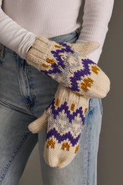 A person stands with arms crossed, wearing Panache Apparel Collection's Cream, Gold, Purple & Gray Patterned Mittens along with a white ribbed long-sleeve top and light blue jeans.