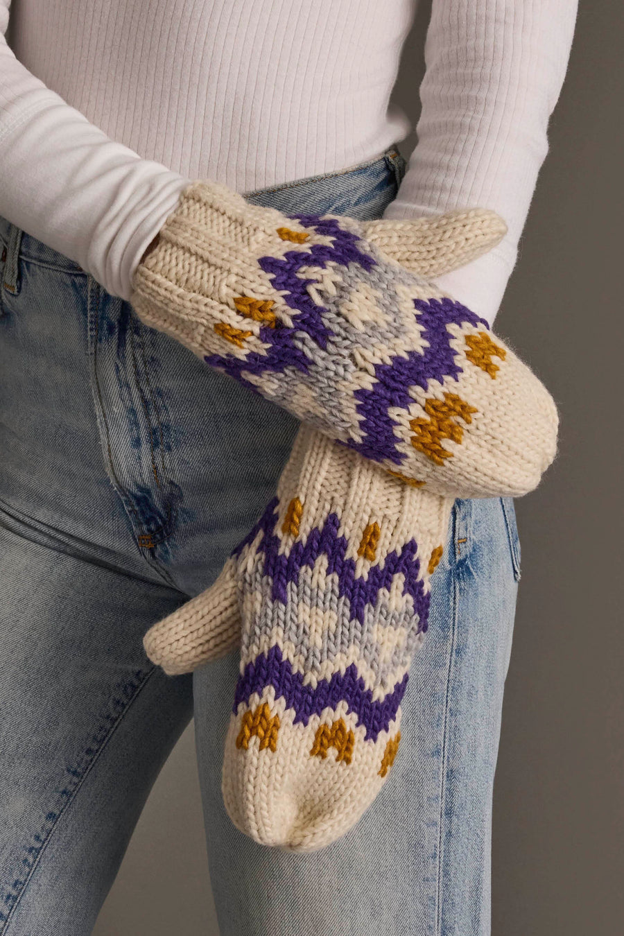 A person stands with arms crossed, wearing Panache Apparel Collection's Cream, Gold, Purple & Gray Patterned Mittens along with a white ribbed long-sleeve top and light blue jeans.