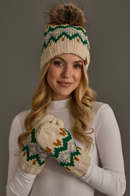 A woman in a white turtleneck wears the Cream, Gold, Green & Gray Patterned Pom Hat from Panache Apparel Collection and matching mittens. She smiles softly against a neutral background.