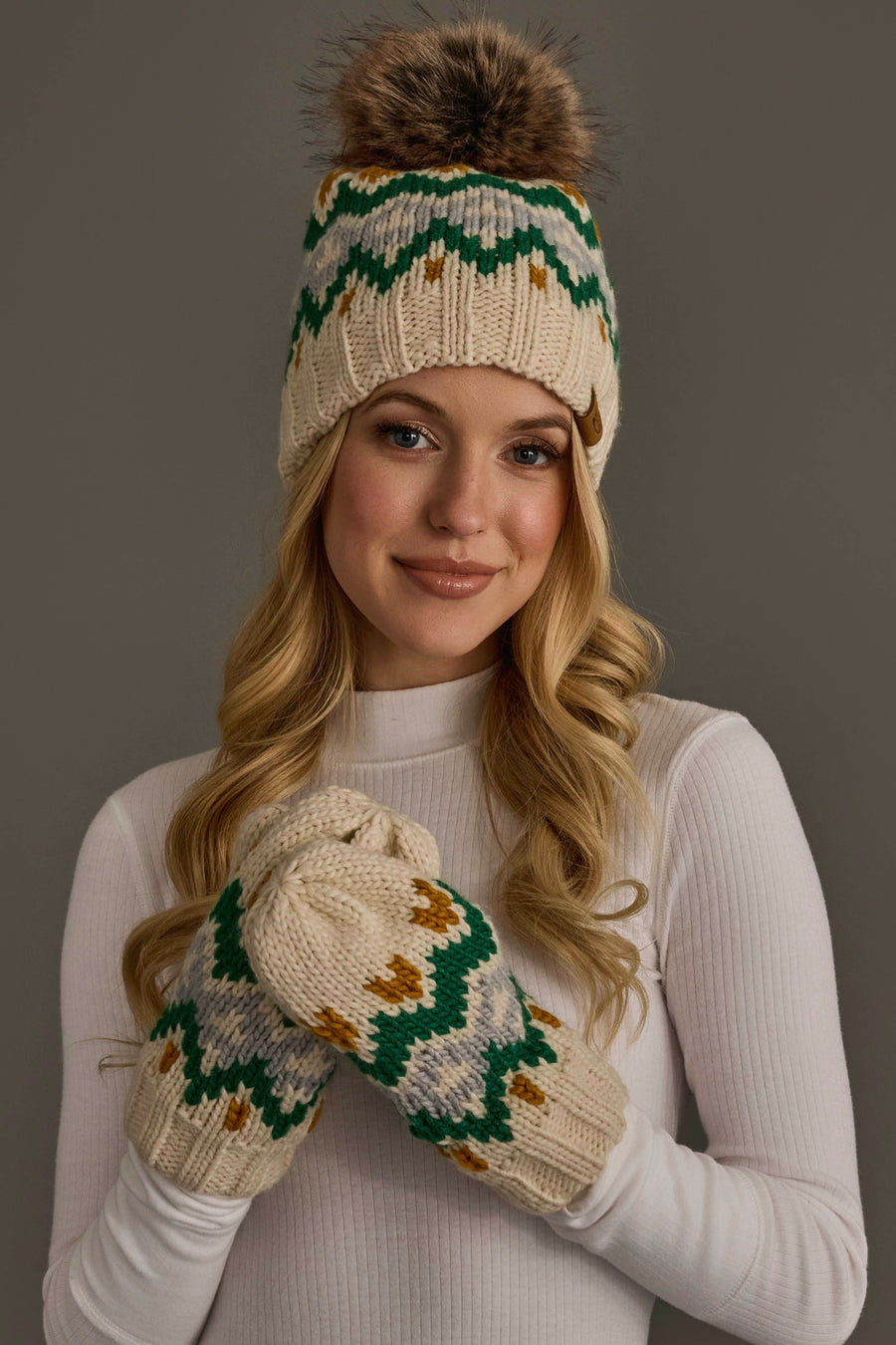 A woman in a white turtleneck wears the Cream, Gold, Green & Gray Patterned Pom Hat from Panache Apparel Collection and matching mittens. She smiles softly against a neutral background.