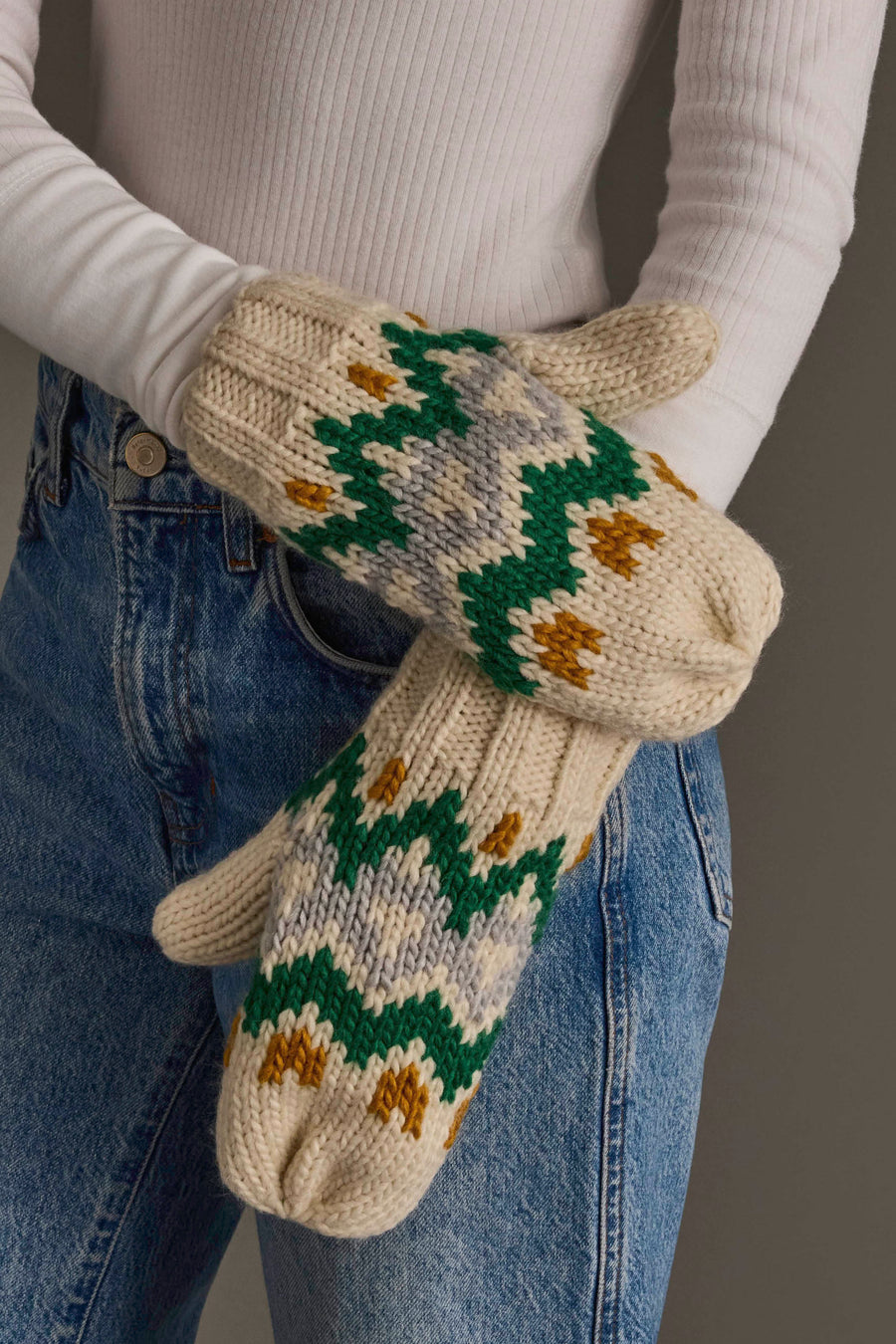A person in a white ribbed shirt and blue jeans holds the Panache Apparel Collection Cream, Gold, Green & Gray Patterned Mittens.