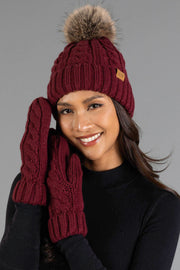 A woman smiles while wearing the Panache Apparel Collection Burgundy Cable Knit Design Pom Hat with a faux fur pom-pom, paired with matching knit mittens and a black sweater, posing against a plain gray background.